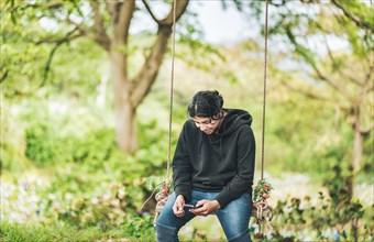 Smiling guy sitting on a swing with smart phone. Young man sitting on a swing using cell phone in