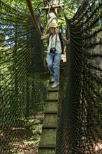 Sporty woman, tourist with camera in treetop path, suspension bridges, chicken ladder, ropes, nets,