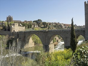 Historic stone bridge with arches over a river, surrounded by a cityscape and vegetation, sunny