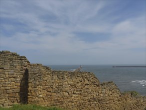 Remains of a wall system near the coast under a blue sky, with a lighthouse in the distance, ruins