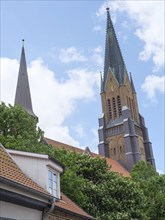 High church tower rises into the blue sky, cloudy, with brick houses and green trees in front,