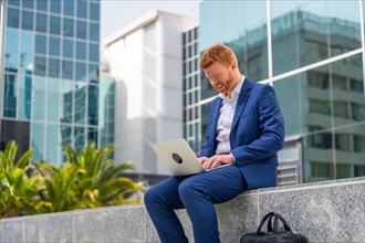 Elegant businessman using laptop sitting outside an office block in the city