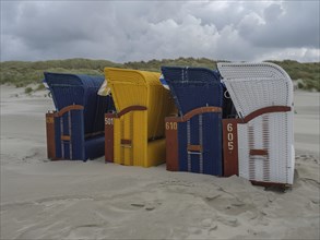 Four colourful beach chairs next to each other on a sandy beach with cloudy sky, colourful beach