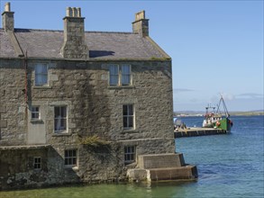 A stone house by the sea with a boat and a jetty in the background, blue sky, old stone house by