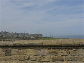 View over stone wall to a row of houses and the sea under a cloudy sky, ruins and old stone walls