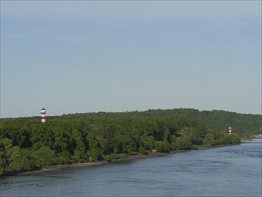 A lighthouse at the edge of a forest, with the river in the foreground under a slightly cloudy sky,
