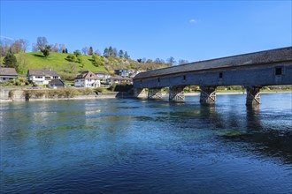Massive wooden bridge over a clear river, surrounded by houses and green hills, under a blue sky,
