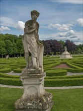 Stone statue of a woman in a well-kept baroque garden with symmetrical hedges and cloudy sky, park