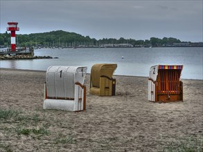 Several beach chairs standing on a cloudy beach, behind them a lighthouse and calm sea under a