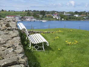 Two white benches at a stone wall, grass in front and a lake with boats and houses behind, partly