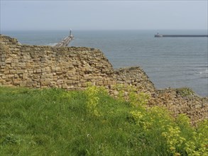 Historic walls rise above green grass with a lighthouse reaching into the sea, ruins and old stone