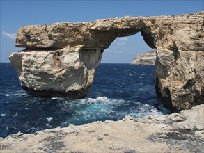 Natural rock arch stretches over the sea, surrounded by coastal cliffs under a blue sky, many