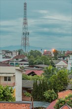 Communication tower above rooftops under cloudy morning sky. Taken from fourth story window in