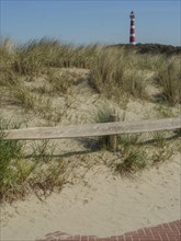 A lighthouse stands behind sandy dunes and grasses with a clear blue sky, red white lighthouse by