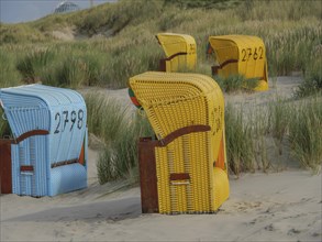Colourful beach chairs standing on the sandy beach, surrounded by green dunes and a cloudy sky,