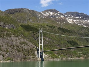 Bridge spanning forested mountains and a green lake, under a clear blue sky, bridge in a fjord with