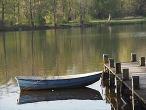 A boat is moored to a jetty and reflected in the calm waters of a lake, rowing boats and pedal