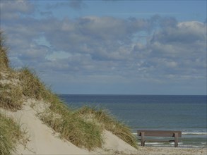 Sand dunes with green grass, behind them the blue sea, a bench and a cloudy sky, dunes on an island