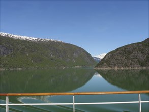 A lake with green mountains and snow-capped peaks seen from a boat with a wooden railing, calm