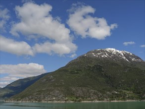 A mountain under a clear blue sky with some clouds, surrounded by a lake and forests, greenish