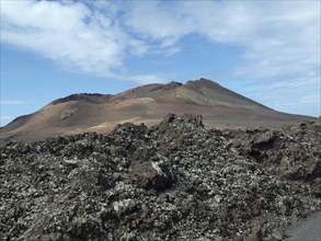 A brown volcano under a partly cloudy sky, surrounded by lava fields and rocks, barren landscape