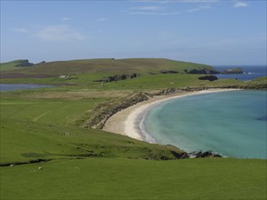 The landscape shows a sandy beach surrounded by green hills and crystal clear blue water under a