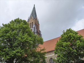 Gothic church tower with pointed spire surrounded by trees against a cloudy sky, large church