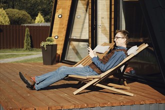 Middle aged man using smartphone while resting on terrace of wooden cabin in a sunny day