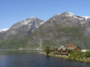 Wooden houses on the riverbank in front of snow-capped mountains under a clear blue sky, spring