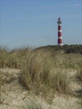 Red and white lighthouse stands between tall dunes with blue sky and few clouds, red and white