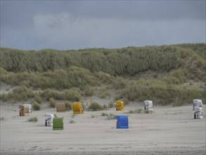 Different coloured beach chairs are spread out in front of a high dune landscape under a cloudy