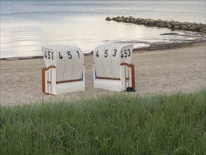 Two white beach chairs in front of a green meadow, sandy beach and calm sea in the background,