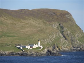 Lighthouse on a rocky coast, surrounded by green hills and blue sea under a clear sky, white