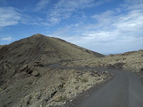 A narrow road winds through a rocky volcanic landscape under a blue sky, barren landscape with