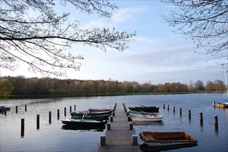 Several boats are moored at a long jetty on a lake, surrounded by spring trees and cloudy skies,