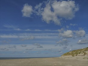Extensive sandy beach under a clear blue sky with scattered clouds, clouds on the beach with dunes