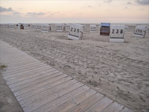 A wooden path leads to empty beach chairs on a quiet beach under a cloudy evening sky, setting sun