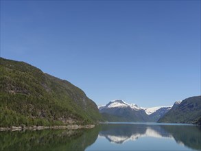 A calm lake, surrounded by snow-capped and forested mountains, under a clear blue sky, calm water