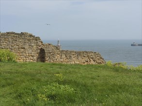 Remains of an old wall in front of a calm sea with a lighthouse in the distance, ruins and old