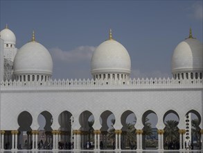 Row of white domes with golden accents and arches, surrounded by palm trees, under a blue sky,