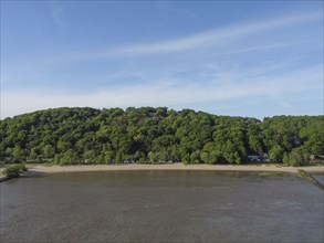 Wide, quiet beach in front of a lush green, wooded hill on the riverbank. Blue sky in the