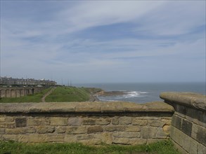 View over the stone wall to the sea and the coast under a cloudy sky with green areas, ruins and