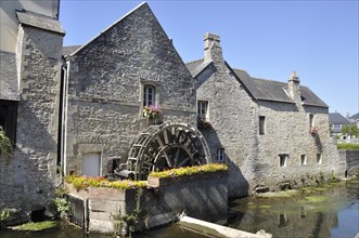 Historic stone buildings with a water wheel, surrounded by flowers and a calm river under a clear