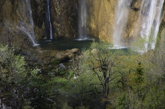 A waterfall cascades down between rocks, surrounded by green foliage and trees. Plitvice Lakes