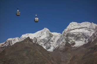 Two cable car gondolas hovering in front of snow covered mountains under a clear blue sky, snow on