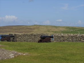 Two historic cannons in front of a stone wall with a view of a hilly landscape and blue sky, old