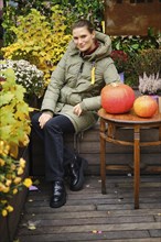 Smiling woman in down jacket sits on bench surrounded with autumn flowers