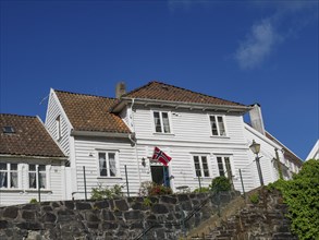 White painted house with red tiled roof, stone wall in the foreground, norwegian flag in front of