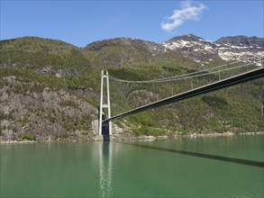 High bridge over a calm lake, surrounded by wooded and rocky hills under a bright blue sky, bridge