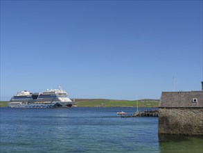 Large cruise ship near the coast with an old building and a small pier in the clear blue water, old
